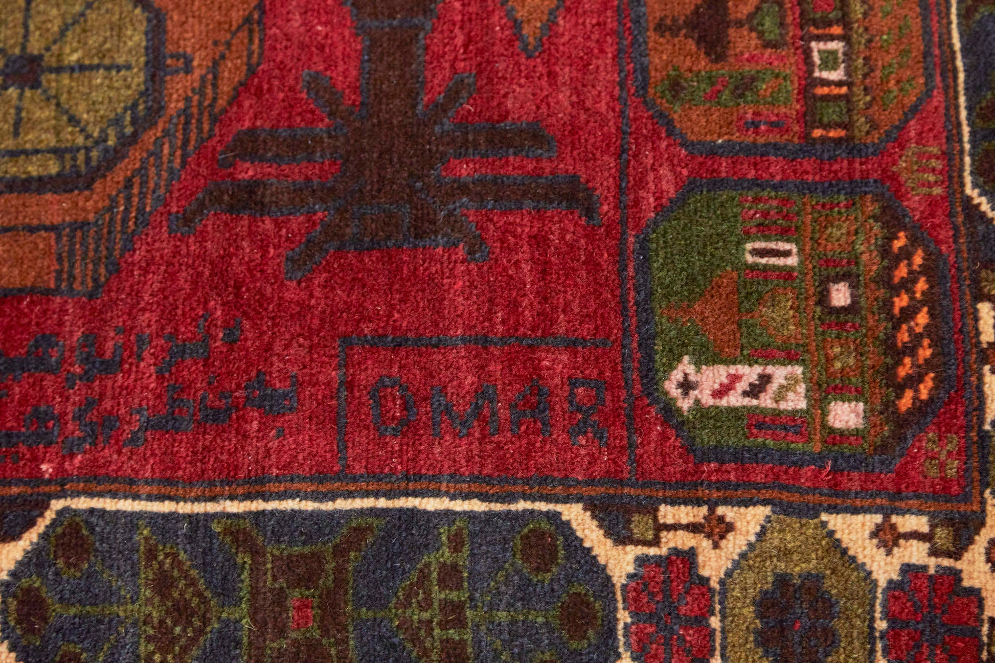 Detail of corner with text "Omar" on Afghan hand woven "Unexploded Ordinance" rug, with deep red base, and green, yellow, tan and cream images depicting weapons, buildings and flowers - Available from King Kennedy Rugs Los Angeles