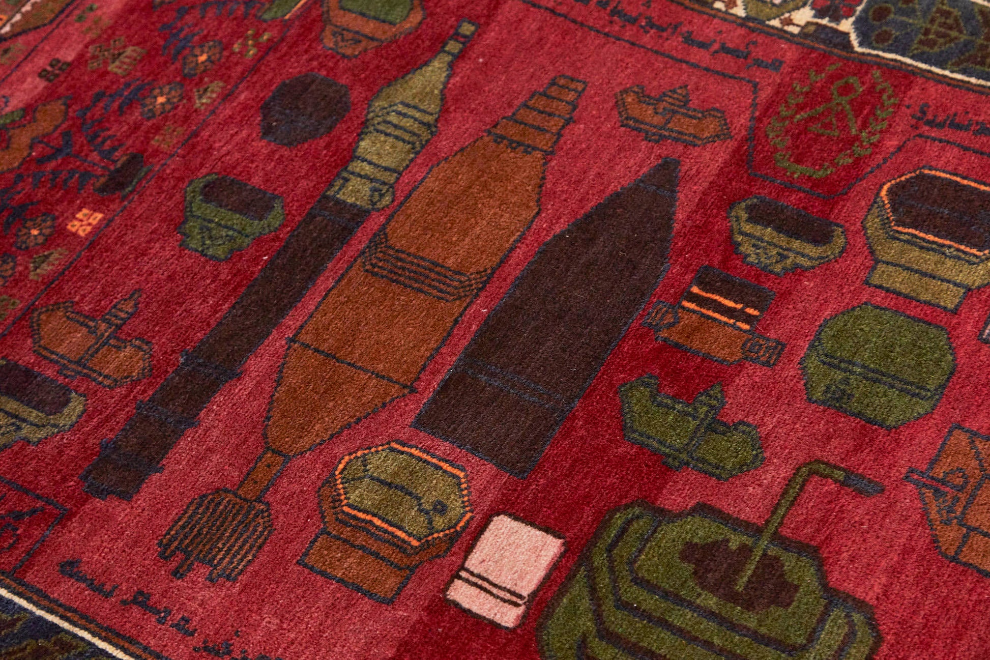 Detail of Afghan hand woven "Unexploded Ordinance" rug, with deep red base, and green, yellow, tan and cream images depicting weapons, buildings and flowers - Available from King Kennedy Rugs Los Angeles