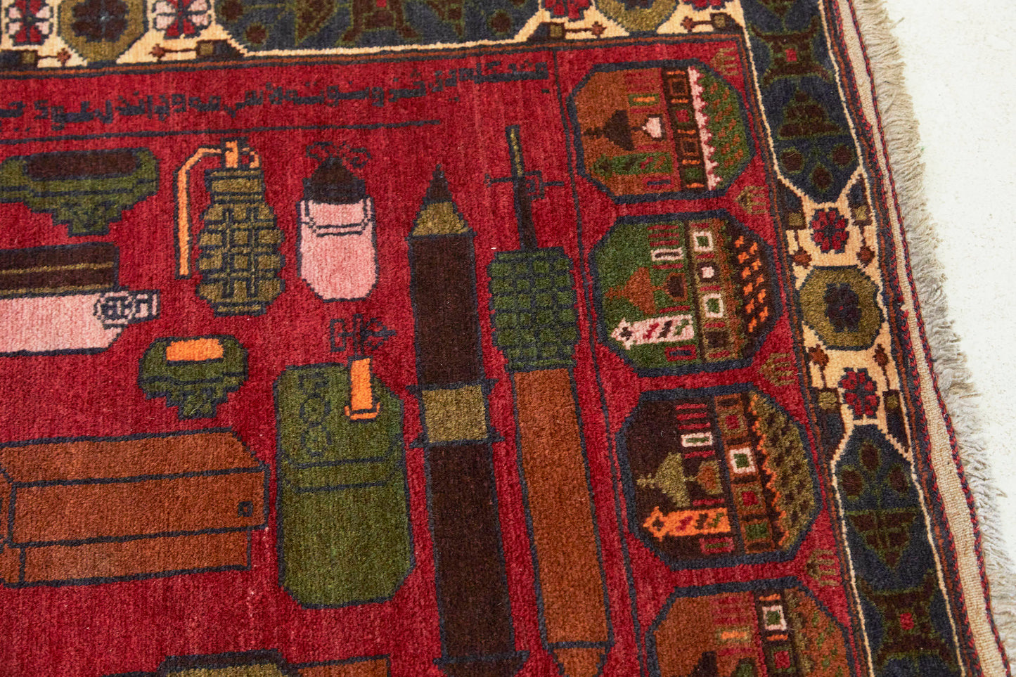 Details of Afghan hand woven "Unexploded Ordinance" rug, with deep red base, and green, yellow, tan and cream images depicting weapons, buildings and flowers - Available from King Kennedy Rugs Los Angeles